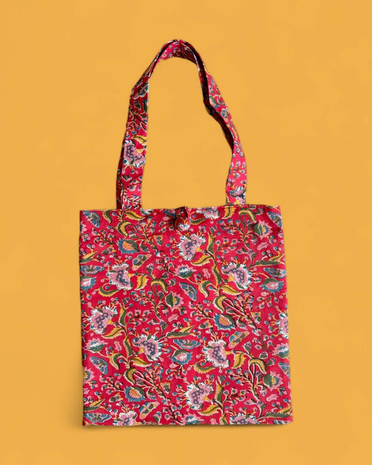 Borsa shopper in cotone stampata a mano con motivo floreale multicolore su fondo rosso, perfetta per l’estate e facile da abbinare.